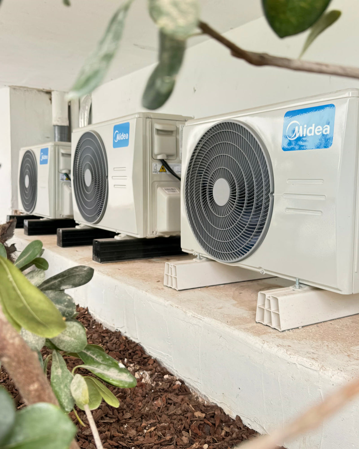 Unidades exteriores de climatización alineadas sobre poyete encalado en el jardín de una villa, entre olivos.