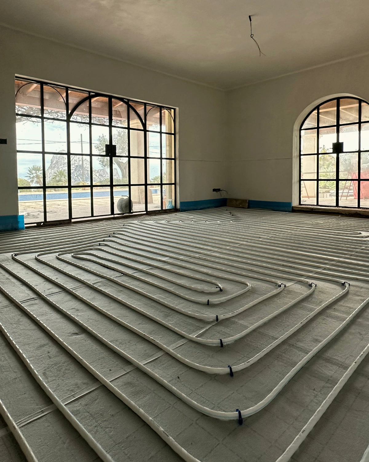 Circuito de suelo radiante tendido sobre panel aislante en el salón de una villa en Ibiza, con ventanales arqueados de acero y vistas al mar.